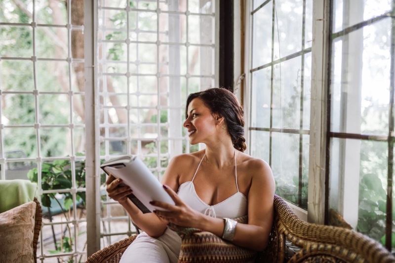 A woman in a summer dress reads a book in a sunroom filled with natural light.