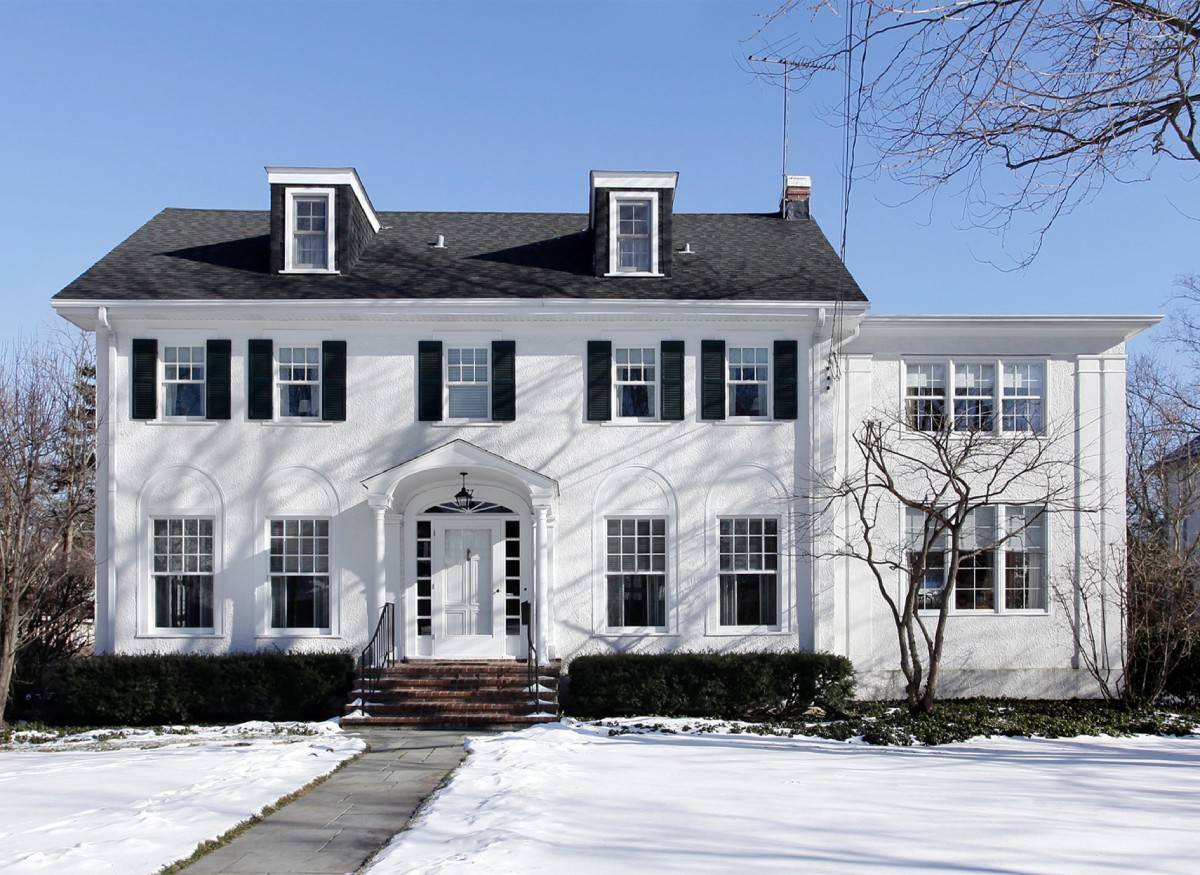 Classic two-story white colonial-style home with black shutters and a symmetrical facade, set against a clear winter sky with snow-covered lawn—showcasing timeless curb appeal and elegant design.